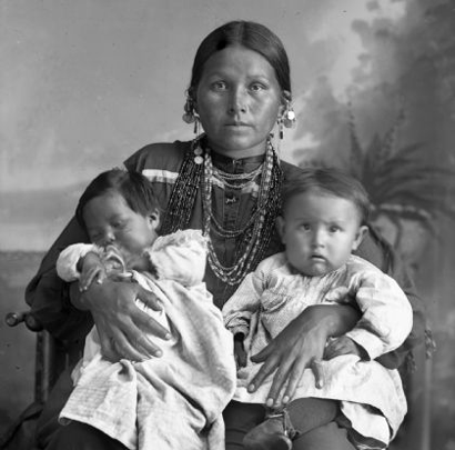 Image description: A black-and-white photo of Mountain Wolf Woman in her traditional clothing and holding one infant on each knee.