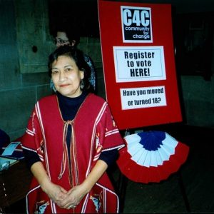 Image description: A photo of Maria Luisa Morales standing in front of a Community for Change sign that encourages people to register to vote.