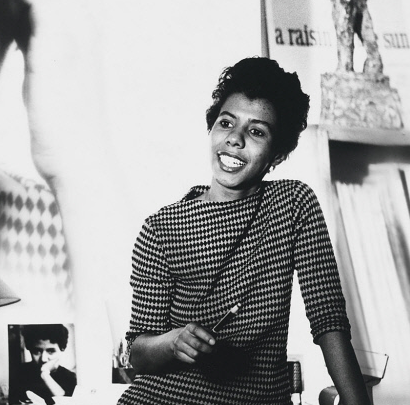 Image description: A black-and-white photo of Lorraine Hansberry holding a pencil with her right hand and using her left to lean on a desk.